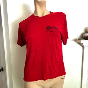 May include: A bright red short-sleeved t-shirt featuring a black graphic on the upper left chest. The graphic includes a stylized logo above the text KITCHEN STAFF. The shirt has a crew neckline and a straight hemline, displayed on a mannequin.