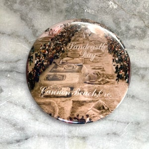 May include: A round, glossy button featuring a sepia-toned image of a sandcastle competition. The text "Sandcastle Day" and "Cannon Beach, Ore." are visible. The image shows numerous sand sculptures and a crowd of people.