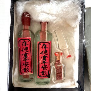 May include: A collection of small, clear glass bottles with red stoppers and labels, presented in a black box with white padding. The labels feature Chinese characters. One bottle is broken. The smallest bottle is filled with small, reddish-brown beads.