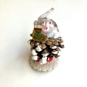 May include: A vintage Christmas ornament featuring a gnome emerging from a pinecone. The gnome has a white hat and holds a green and red stocking. The pinecone is painted with white and red accents, and sits on a silver base.