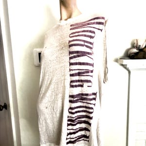 May include: A light-colored t-shirt with the word "WORKING" printed vertically in a dark maroon color. The shirt is displayed on a mannequin. The fabric appears to be lightweight and slightly textured.