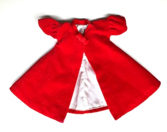 May include: A bright red doll-sized cape with short sleeves and a white satin lining. The cape has a small bow at the neckline and is open in the front. The garment is likely for a doll or other small toy.