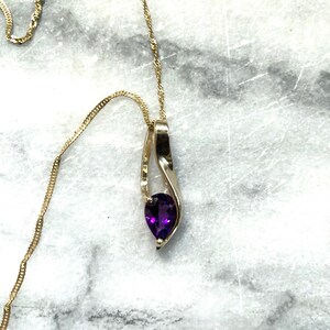 May include: A gold-toned necklace with a teardrop-shaped pendant. The pendant features a vibrant purple gemstone set within a curved, open-frame design. The chain is delicate and twisted, and the necklace is displayed against a marbled background.