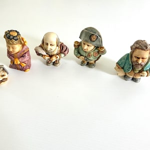 May include: A collection of miniature figurines depicting historical figures. The figurines include a scholar, William Shakespeare, Napoleon Bonaparte, and a bearded man. Each figure is painted with detailed clothing and facial features, and they are arranged on a white surface.