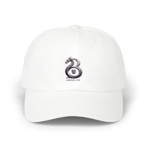 Peut inclure: Une casquette de baseball blanche arborant un motif central représentant la lettre B stylisée en forme de dragon, avec un petit écusson à l'intérieur de la courbe et le texte BRSPUK LTD imprimé en petites lettres noires en dessous.