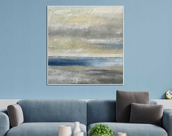 May include: A blue sofa with grey cushions sits in front of a light blue wall with a framed abstract painting of a seascape in shades of grey, blue, and beige. A wooden coffee table with a black metal frame sits in front of the sofa.