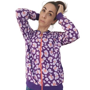 Könnte beinhalten: Eine langärmelige Reißverschlussjacke mit einem Leopardenmuster in Violett, Hellrosa und Pfirsich. Das Kleidungsstück verfügt über einen leuchtend orangefarbenen vertikalen Reißverschluss, einen Rundhalsausschnitt sowie violette Rippbündchen.