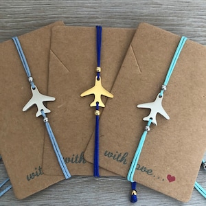 May include: Three friendship bracelets with gold airplane charms and blue, light blue, and turquoise cords. The bracelets are tied with a bow and have the words "with love..." on the brown card.