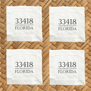 May include: Four white square coasters with stitched edges. Each coaster features the text "33418 PALM BEACH GARDENS FLORIDA" in black, centered within a stitched border. The coasters are arranged on a woven surface.