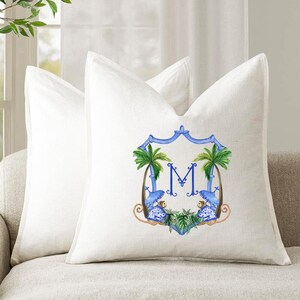 May include: Two white linen throw pillows on a beige sofa. One pillow features a blue and white crest design with the letter "M", palm trees, and monkeys. The other pillow is plain white.