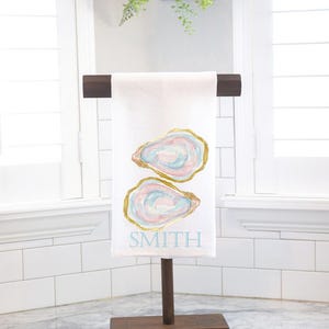 May include: White kitchen towel with two oyster shell designs in pastel colors and gold trim. The word "SMITH" is printed in light blue below the oyster shells. The towel is displayed on a wooden stand.