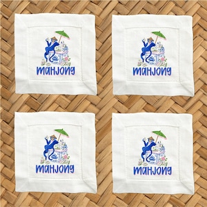 May include: Four white square cocktail napkins with embroidered designs. Each napkin features a blue monkey with a green umbrella, a blue and white pagoda, and the word "MAHJONG" in blue script. The napkins are on a woven background.