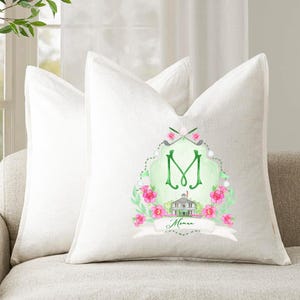 May include: Two off-white linen throw pillows on a beige sofa. One pillow features a watercolor design with a green letter "M", pink flowers, golf clubs, and the word "Moman".