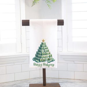 May include: White hand towel with a Christmas tree design made of green Mahjong tiles, topped with a gold star. The words "Merry Mahjong" are printed below the tree. The towel is displayed on a wooden stand.