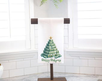 Könnte beinhalten: Weißes Handtuch mit einem Weihnachtsbaum-Design aus grünen Mahjong-Steinen, gekrönt von einem goldenen Stern. Die Worte "Merry Mahjong" sind darunter gedruckt. Das Handtuch wird auf einem Holzständer präsentiert.