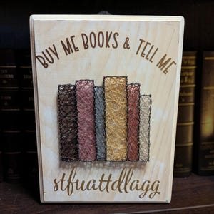 May include: A rectangular wooden plaque featuring string art of six books in brown, pink, blue, yellow, and cream colors. The text "BUY ME BOOKS & TELL ME" is printed in brown at the top, and "stfuattdlagg" is printed in brown at the bottom.