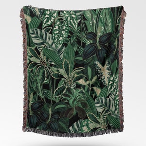 Woven cotton throw blanket spread open to see the full design of botanical plants in a jungle like design. It has many green hues and leaf types. Design is flushed to the border, which has a fringe all around.