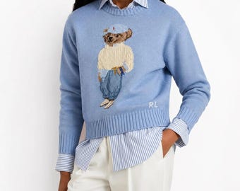 Könnte beinhalten: Ein hellblauer, langärmeliger Strickpullover mit einem mittig aufgestickten Bären, der einen cremefarbenen Zopfmusterpullover, eine blaue Mütze und eine blaue Hose trägt. Die Buchstaben RL sind weiß am Saum eingestickt. Über einem blau-weiß gestreiften Hemd.