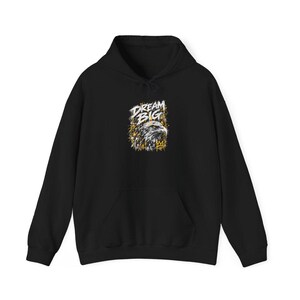 K&ouml;nnte beinhalten: Ein schwarzer Kapuzenpullover mit einer Grafik auf der Brust. Das Design zeigt einen detaillierten Adlerkopf in Wei&szlig;- und Graut&ouml;nen, begleitet vom Schriftzug "DREAM BIG" in wei&szlig;en Buchstaben und gelben Farbspritzern.