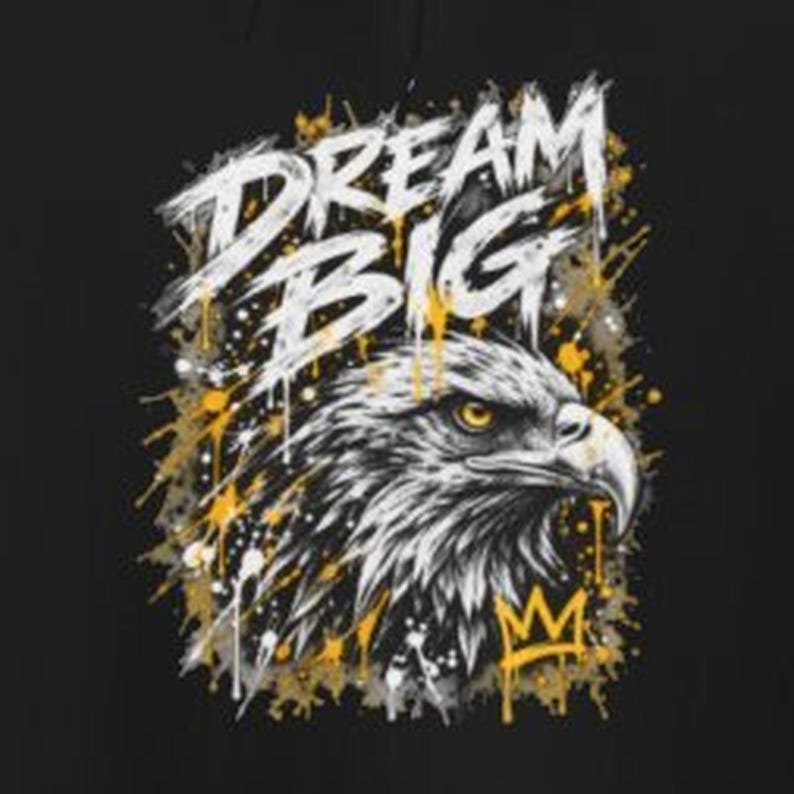 K&ouml;nnte beinhalten: Ein schwarzer Grafikdruck mit einem wei&szlig;en Adlerkopf mit gelben Augen, einem kleinen gelben Kronensymbol und dem Text DREAM BIG in wei&szlig;er, abgenutzter Schrift. Das Design ist von gelben und wei&szlig;en Farbspritzern umgeben.