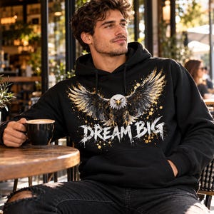 K&ouml;nnte beinhalten: Ein schwarzer Kapuzenpullover mit einem Grafikdruck eines Wei&szlig;kopfseeadlers mit ausgebreiteten Fl&uuml;geln und goldenen Farbspritzern. Der Text "DREAM BIG" ist in einer verwaschenen wei&szlig;en Schrift unter dem Adler auf der Vorderseite gedruckt.
