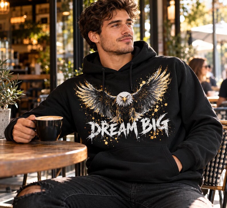 K&ouml;nnte beinhalten: Ein schwarzer Kapuzenpullover mit einem Grafikdruck eines Wei&szlig;kopfseeadlers mit ausgebreiteten Fl&uuml;geln und goldenen Farbspritzern. Der Text "DREAM BIG" ist in einer verwaschenen wei&szlig;en Schrift unter dem Adler auf der Vorderseite gedruckt.