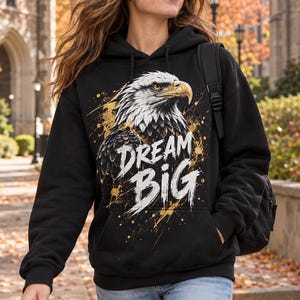 K&ouml;nnte beinhalten: Ein schwarzer Kapuzenpullover mit einer Grafik eines Wei&szlig;kopfseeadlerkopfes im Profil mit wei&szlig;en und braunen Federn. Der Text DREAM BIG ist in Wei&szlig; unter dem Adler aufgedruckt, akzentuiert durch goldene Farbspritzer auf der Brust.