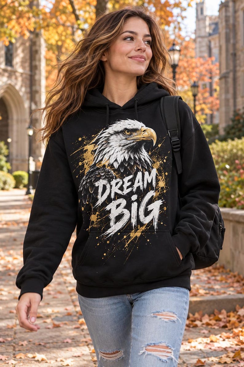 K&ouml;nnte beinhalten: Ein schwarzer Kapuzenpullover mit einer Grafik eines Wei&szlig;kopfseeadlerkopfes im Profil mit wei&szlig;en und braunen Federn. Der Text DREAM BIG ist in Wei&szlig; unter dem Adler aufgedruckt, akzentuiert durch goldene Farbspritzer auf der Brust.