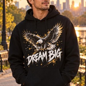 K&ouml;nnte beinhalten: Ein schwarzer Kapuzenpullover mit einem Grafikdruck eines Wei&szlig;kopfseeadlers im Flug mit ausgebreiteten Fl&uuml;geln. Unter dem Adler ist der Text "DREAM BIG" in einer verwaschenen wei&szlig;en Schrift gedruckt, akzentuiert durch gelbe und wei&szlig;e Farbspritzer.