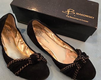 May include: A pair of black suede ballet flats with gold chain embellishments and a bow detail. The shoes are on a light blue surface and have the brand name "B. Makowsky The Signature of Luxury" on the box.