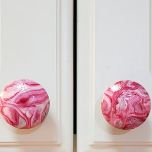 May include: Two pink and white marbled cabinet knobs. The knobs are round and have a swirl pattern.