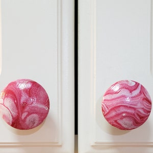 May include: Two pink and white marbled cabinet knobs. The knobs are round and have a glossy finish.