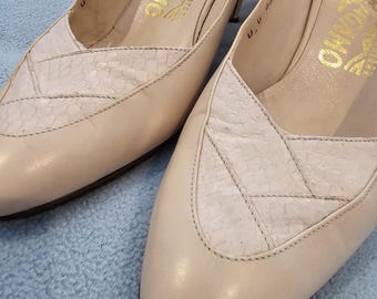 May include: A pair of cream-colored Salvatore Ferragamo slingback heels. The shoes feature a pointed toe and a textured, light-colored panel on the vamp. The heels have a low, stiletto design. The brand name is visible in gold lettering.