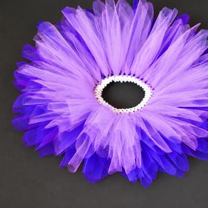 May include: A circular tutu skirt constructed from layers of sheer tulle fabric in alternating shades of light lavender and deep violet. The skirt features a white elasticized waistband that gathers the fabric into a full, ruffled silhouette.