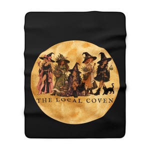 May include: A black fleece blanket featuring a large, circular, golden moon graphic. Inside the moon are five illustrated figures wearing witch hats and outfits, accompanied by a black cat. The text THE LOCAL COVEN is printed in black.
