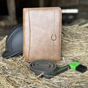 May include: A tan leather-like portfolio featuring a horseshoe icon and the text THE HORSE LIFE, accompanied by a black equestrian helmet, a circular black rubber curry comb, and a green and black hoof pick with a brush.