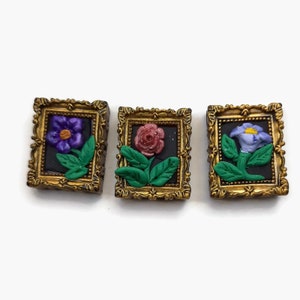 May include: Three miniature framed pictures with gold frames and black backgrounds. Each picture features a different flower: a purple flower, a pink rose, and a blue flower.
