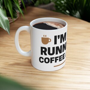 Peut inclure: Une tasse en céramique blanche remplie de café noir. La tasse présente une icône de tasse à café marron et un texte noir en gras indiquant I'M RUNNING ON COFFEE AND DREAMS, avec une ligne horizontale marron sous le texte sur le côté.