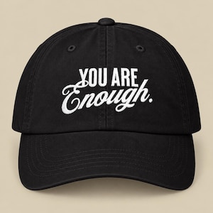 Pode incluir: Um boné de beisebol preto com o texto bordado em branco "YOU ARE Enough." centralizado no painel frontal. O boné possui uma copa estruturada, aba curva e ilhós costurados para ventilação, apresentando um design simples.