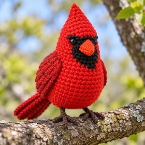 Op de afbeelding: Een gehaakt kardinaalvogel-figuurtje met een felrood lichaam, een zwart masker rond de ogen en een oranje snavel. De knuffelvogel heeft pootjes van donkerbruin garen en is gemaakt met een gestructureerd patroon van gehaakte steken.