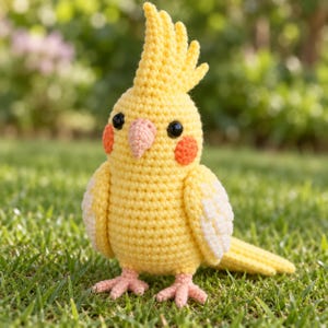 May include: A crocheted cockatiel bird figurine featuring a bright yellow body, a tall crest, white wing accents, and round orange cheek patches. The bird has small black bead eyes and a light pink beak, with matching pink feet.