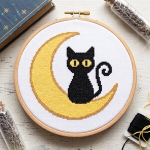 May include: A cross-stitch embroidery piece in a round wooden hoop featuring a black cat with yellow eyes sitting on a yellow crescent moon against a white fabric background. The design is centred within the circular frame of the embroidery hoop.