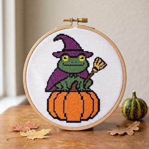 May include: A cross-stitch embroidery piece in a round wooden hoop featuring a green frog wearing a purple witch hat and cape, holding a small broomstick while sitting atop a large orange pumpkin on a white fabric background.