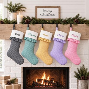Puede incluir: Cinco calcetines navideños colgantes con patrones de cuadros vichy en negro, verde azulado, amarillo, morado y rosa. Cada calcetín tiene un puño blanco con borde de volantes y un nombre bordado en letra cursiva: Dad, William, Mom, Maria y Olivia.