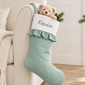 Puede incluir: Un calcetín navideño de cuadros vichy verdes y blancos con un puño blanco bordado con el nombre Kevin en letra verde. Un oso de peluche marrón claro con pajarita de cuadros asoma por el calcetín junto a unas ramas de pino.