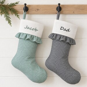 May include: Two Christmas stockings hanging from black hooks. One stocking features a green gingham pattern with the name Jacob embroidered on a white cuff. The other stocking has a black gingham pattern with the name Dad on a white cuff.