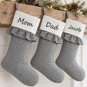 May include: Three Christmas stockings made of black and white gingham fabric with a ruffled cuff. Each stocking features a white top band with the names Mom, Dad, and Jacob printed in black script lettering, hanging from a wooden mantel.