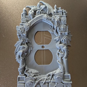 May include: A gray, decorative light switch cover featuring a fantasy castle design with dragons. The cover has two circular openings for outlets and is adorned with detailed relief sculptures of dragons, turrets, and shields.