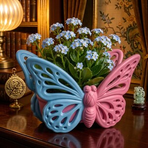 May include: A ceramic planter shaped like a butterfly with a light blue left wing and a pink right wing, featuring cut-out details. The planter holds a cluster of small, light blue flowers with green stems and leaves, creating a vibrant display.