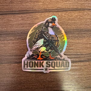 May include: A holographic sticker featuring a white goose dressed in tactical military gear, including a helmet, vest, and rifle. The goose stands on a dark mound against a circular background. The text HONK SQUAD is printed in bold letters at the bottom.